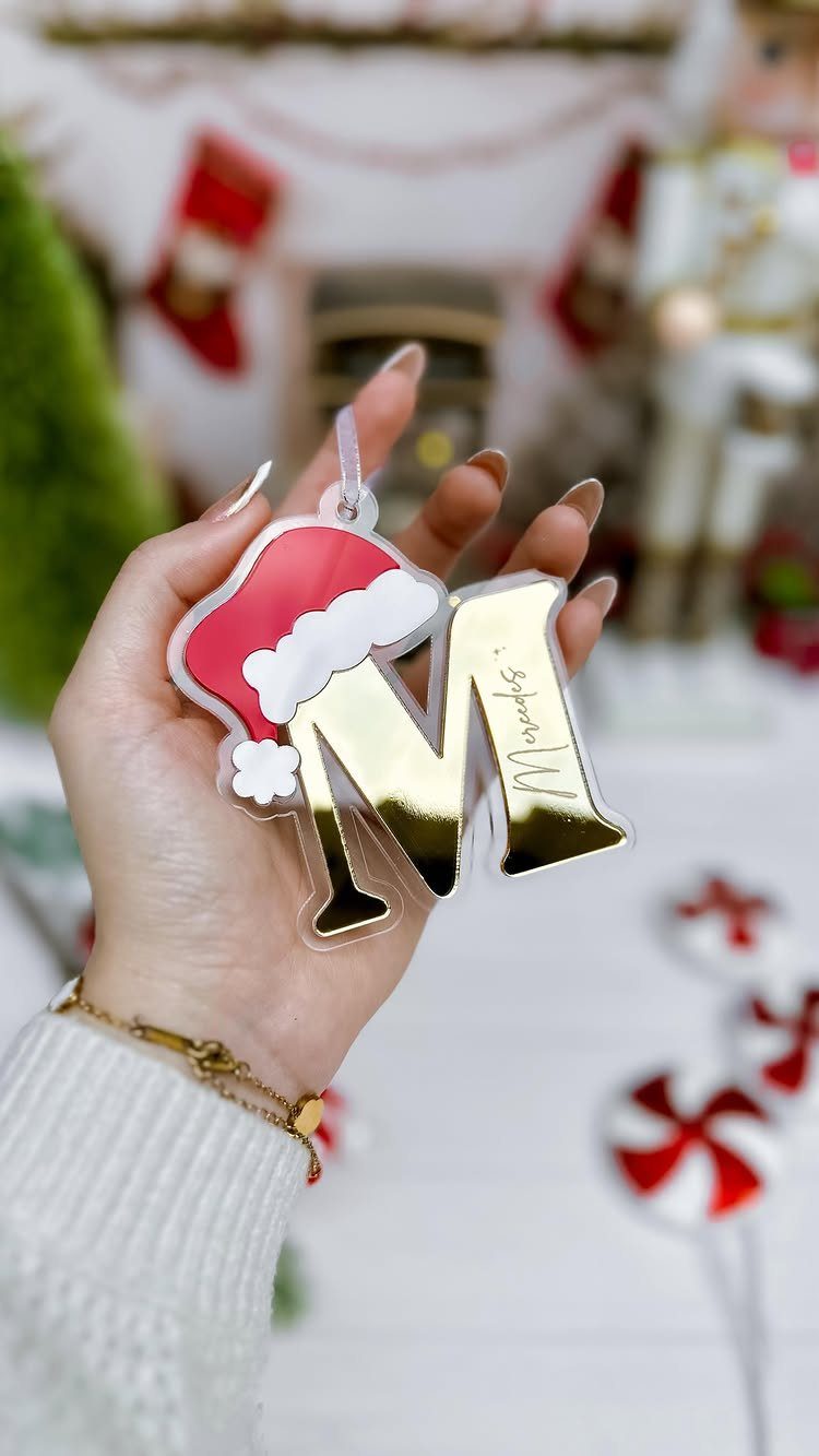 A hand holding a gold letter M ornament decorated with a small red Santa hat.