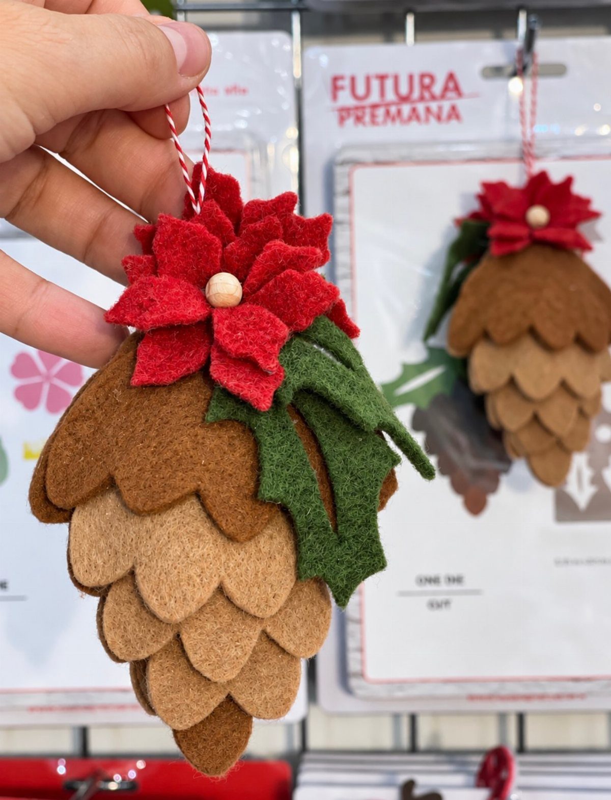 A hand holding a brown felt pinecone ornament decorated with a red poinsettia and green leaves.