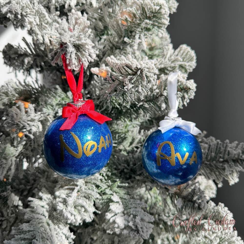 Two blue glittery ornaments with gold names hanging on a snowy Christmas tree branch.