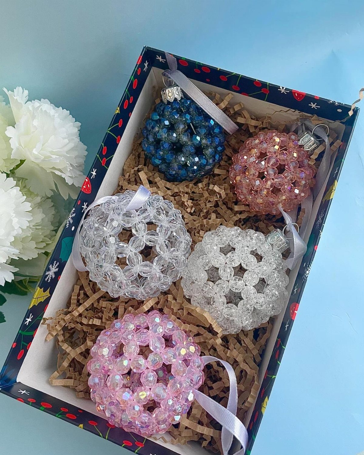 Five sparkling beaded Christmas ornaments in blue, pink, and clear resting in a gift box.