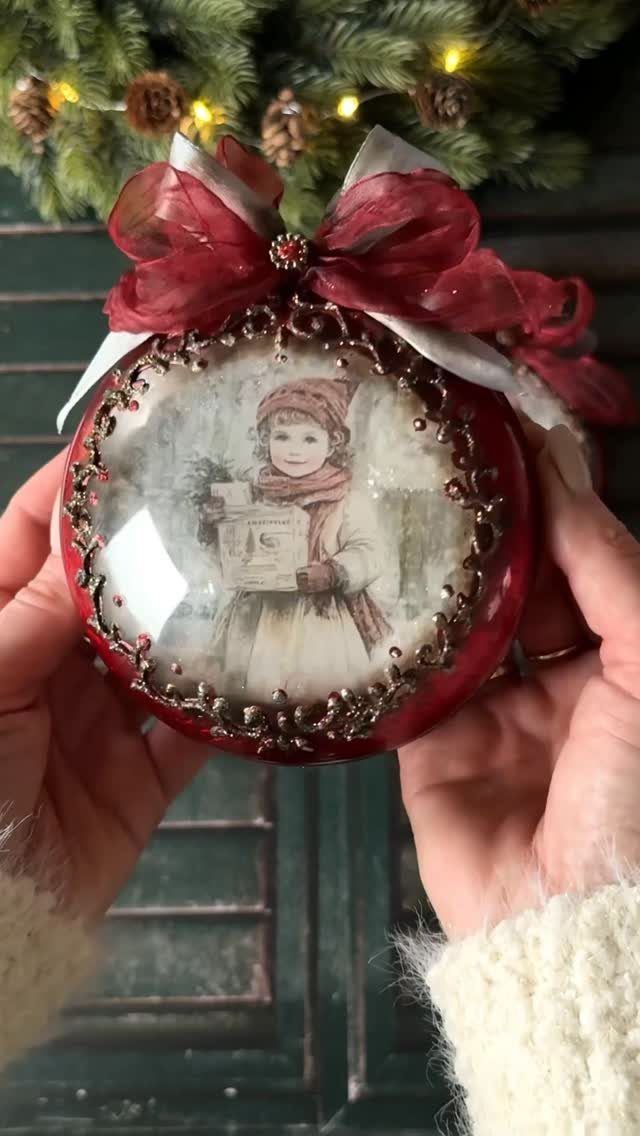 Hands holding a red vintage-style decoupage Christmas ornament with a large red bow.