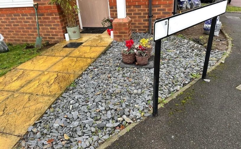 Grey slate chips cover a garden bed containing three small flower baskets near a yellow walkway.