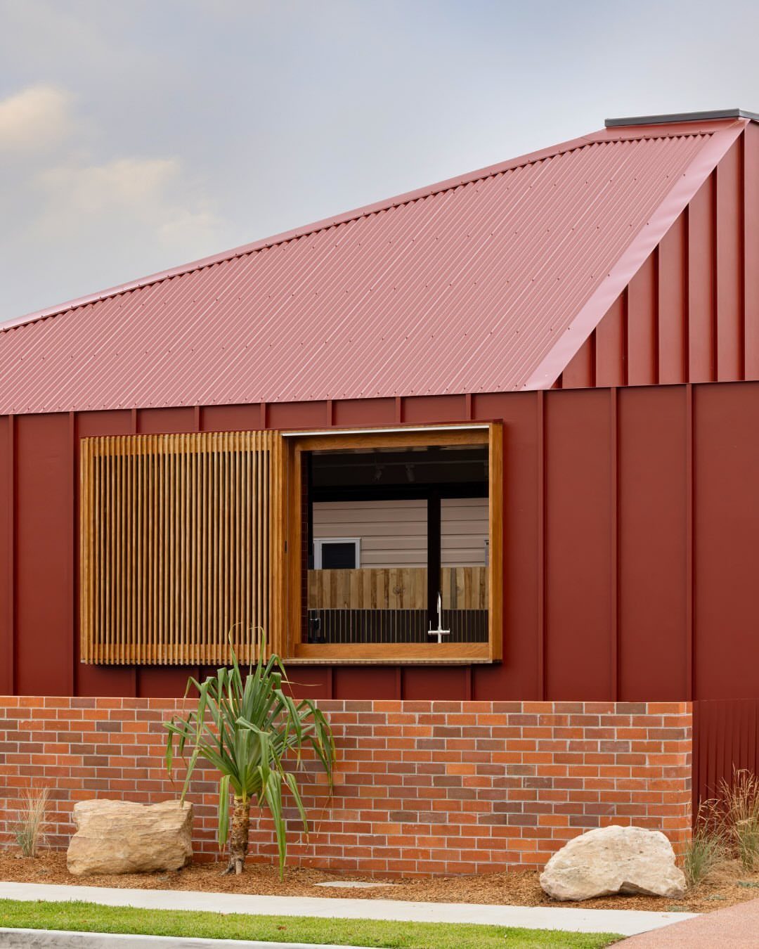 Burgundy red vertical metal siding on a modern building with a brick base and wooden window slats.