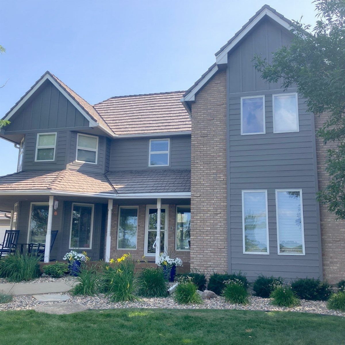 A two-story house with dark gray siding, tan brick, and white window trim surrounded by green plants.