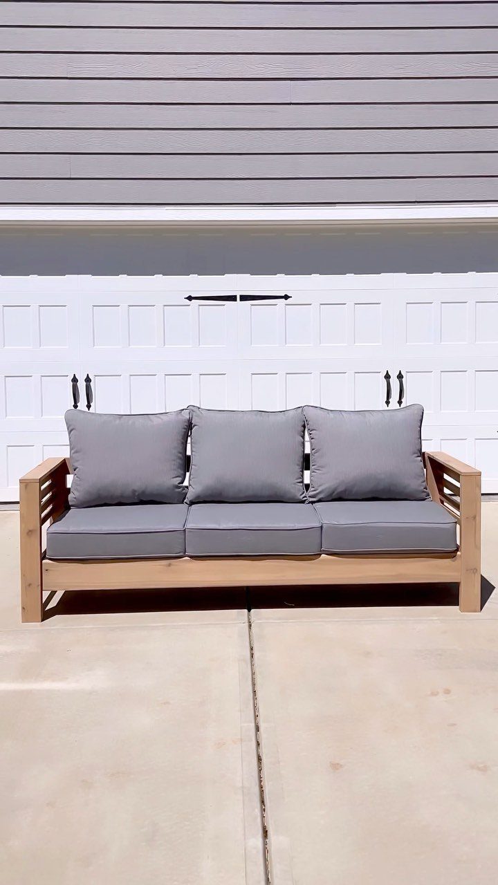 A wooden outdoor couch with grey cushions and pillows sitting on a concrete driveway.