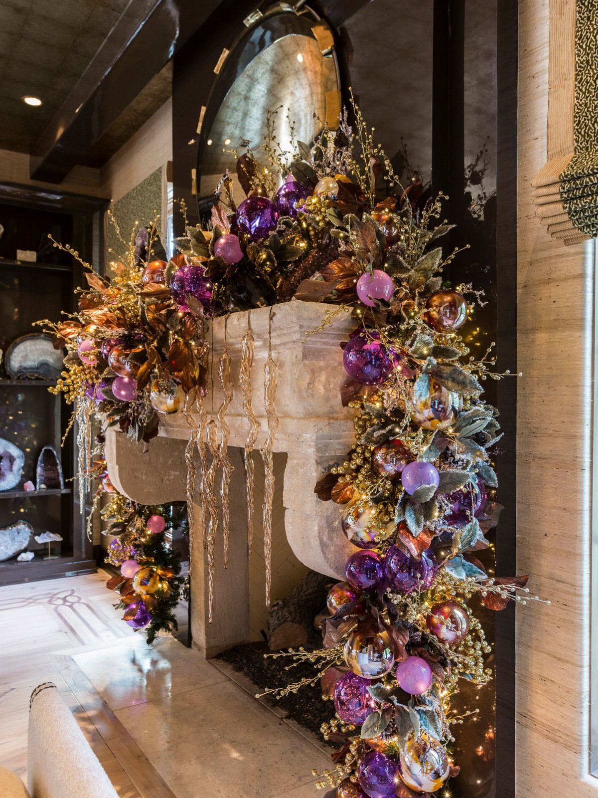 Ornate purple and gold Christmas decorations cascading down a stone fireplace mantel in a luxury home.