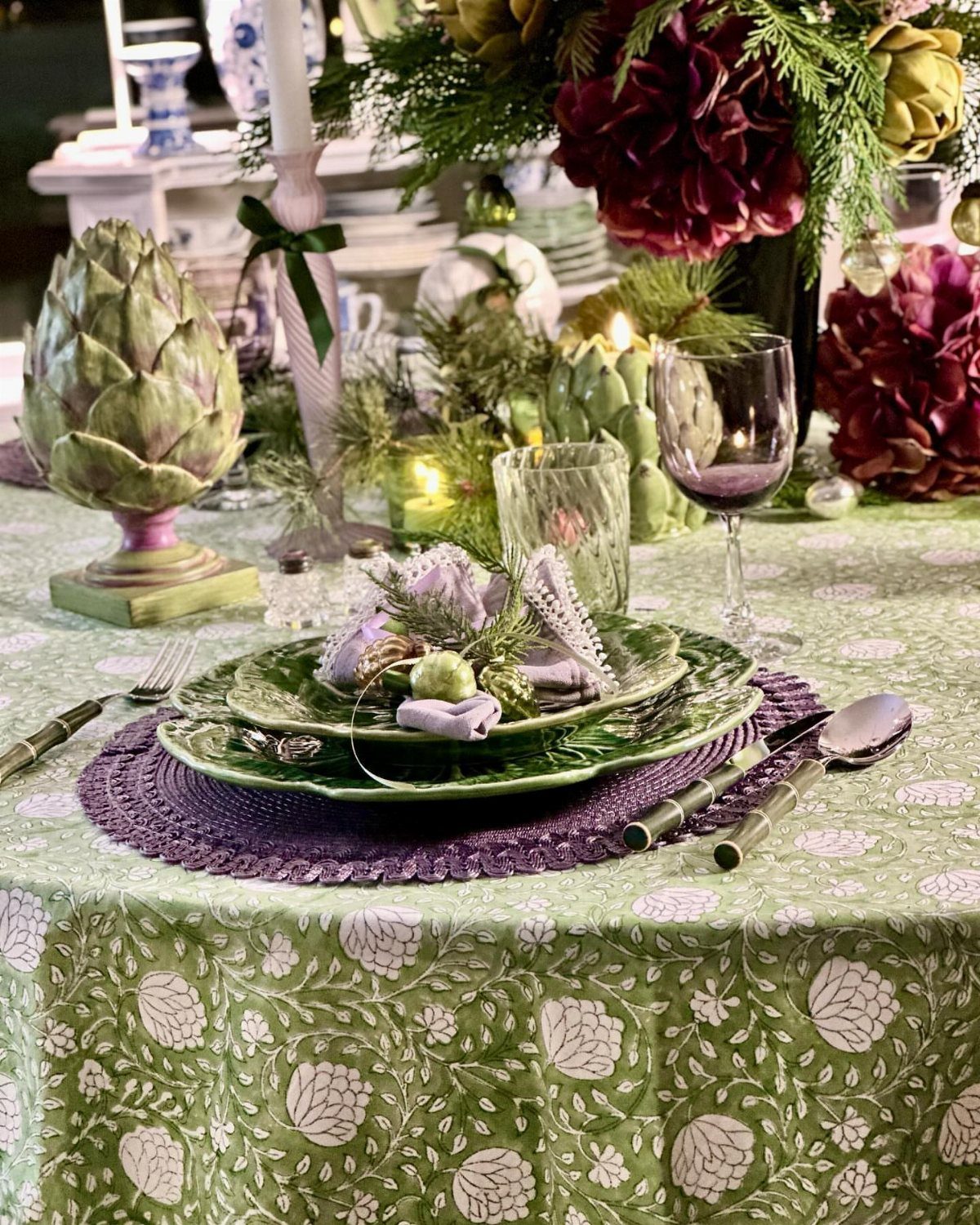A festive table setting with green patterned cloth, purple placemats, green plates, and floral centerpieces.
