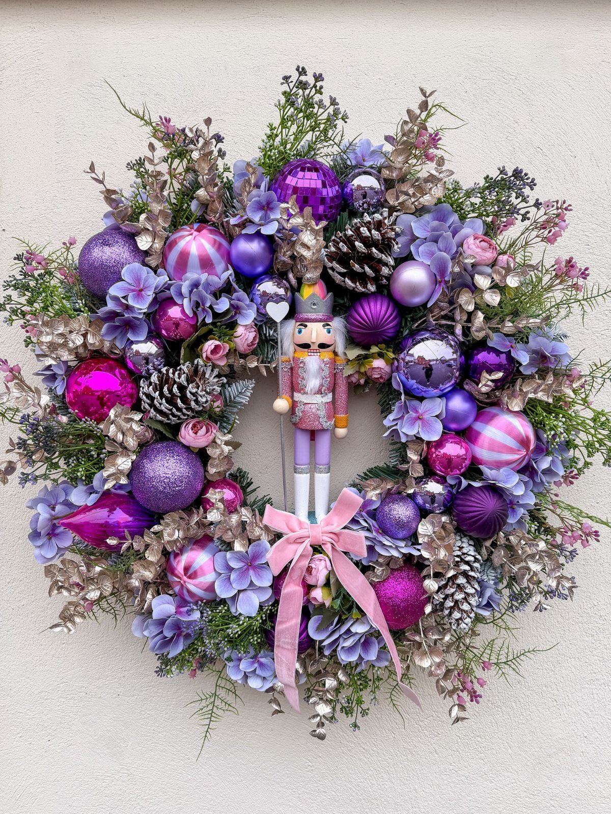 A festive purple Christmas wreath featuring ornaments, flowers, pinecones, and a small nutcracker figurine.