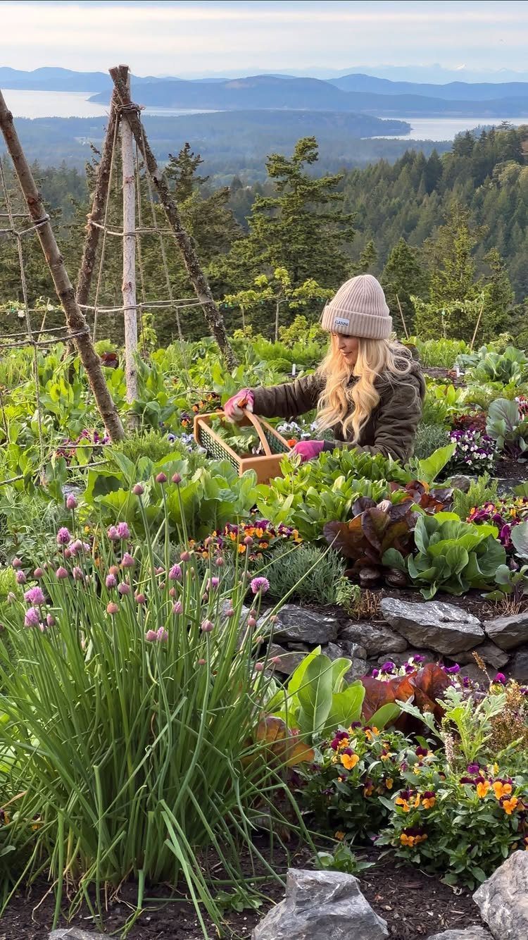 A woman gardening among leafy greens, chives, and flowers with a mountain landscape in the background.